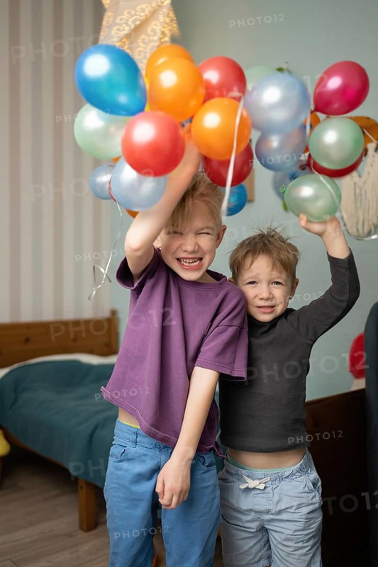 Two joyful children play indoors, holding several colorful balloons. Their happy expressions capture the excitement and fun of childhood celebrations and indoor playtime