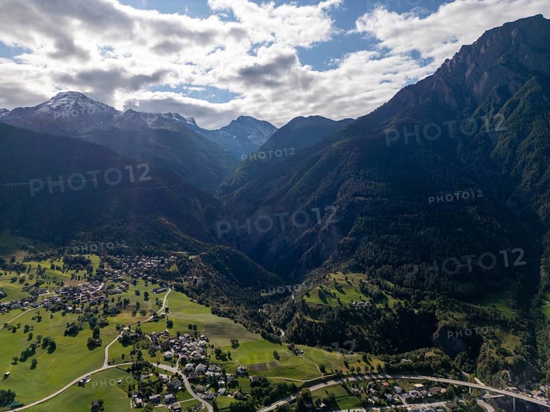 A breathtaking aerial view of the Brig area in Switzerland, showcasing lush green valleys, scattered villages, and the majestic Alps under a partly cloudy sky