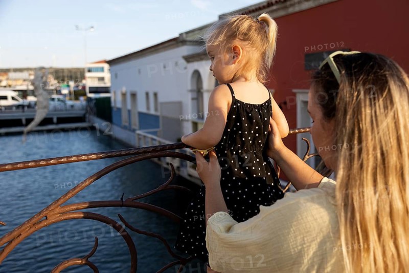 A mother lovingly holds her young daughter as they enjoy a sunlit day outdoors by a serene waterfront. The child wears a polka dot dress, embracing the warm atmosphere