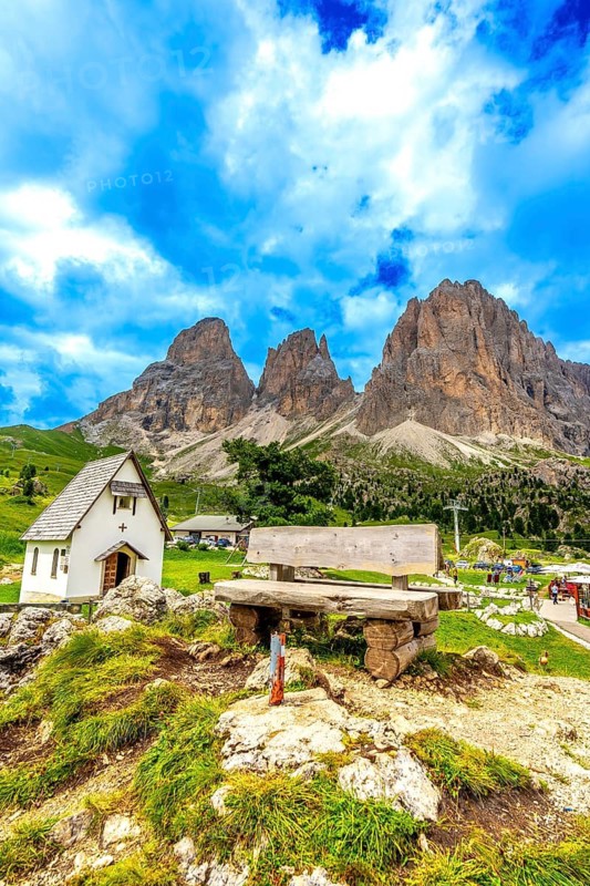 Picturesque landscape showcasing a wooden bench, a small chapel, and the towering peaks of the sella group in the italian dolomites, under a vibrant summer sky