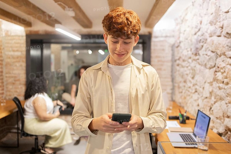A young man with red hair is focused on his smartphone in a trendy coworking space, while others work in the background. A laptop sits on the wooden desk nearby