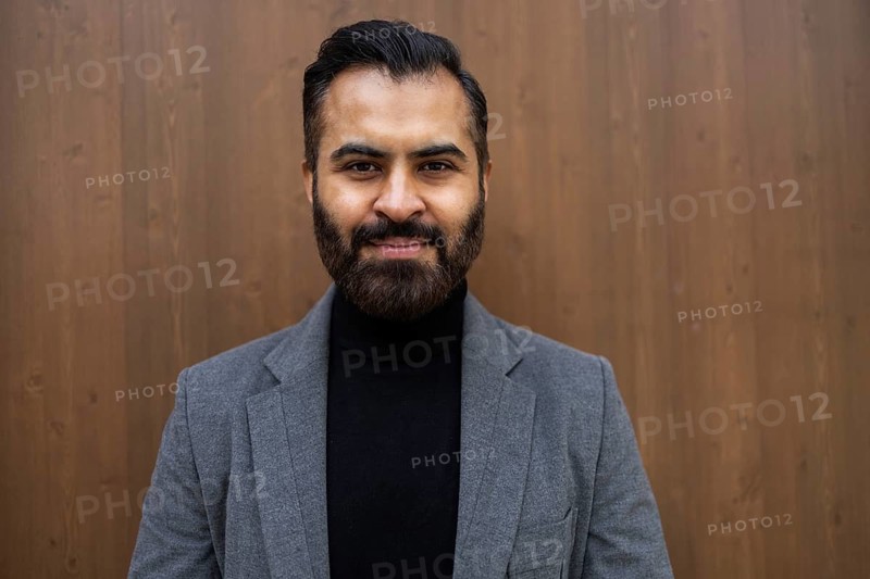 A well-dressed Indian man in business attire stands confidently against a wooden background in his winter outfit, consisting of a gray coat and a black turtleneck, looking directly at the camera