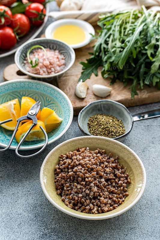 A vibrant display of healthy lunch ingredients includes fresh rocket, oregano, cherry tomatoes, Himalayan salt, buckwheat, lemon, and organic olive oil