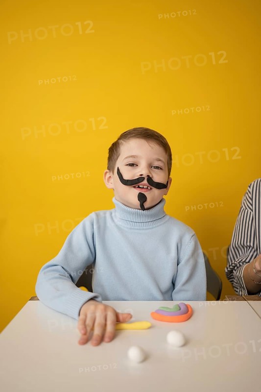 A boy with a playful clay mustache engages in creative activities at a white table, surrounded by colorful modeling clay, against a bright yellow background, inspiring creativity