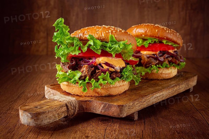 Two burgers are stacked high with fresh lettuce, tomato slices, grilled onions, and a savory sauce. They sit on a wooden board in a warm light setting. Ideal for a meal or snack