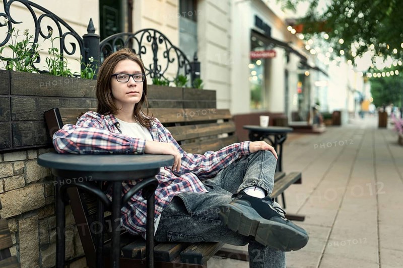 A teenage boy with long hair and glasses sits casually in an outdoor cafe setting, wearing a plaid shirt and jeans, conveying a relaxed, youthful vibe in an urban environment