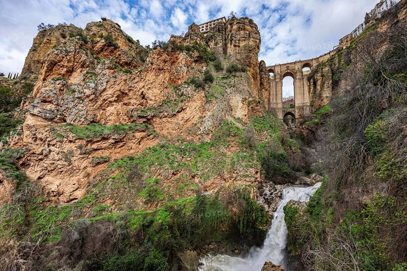 Wide angle view from the bottom of the gorge featuring the massive arches of Puente Nuevo bridge and a small waterfall in Ronda, Spain