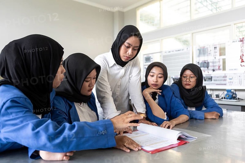 Asian Muslim teacher instructing a group of attentive students in a classroom setting. The students, wearing headscarves, focus intently on a textbook as the teacher explains