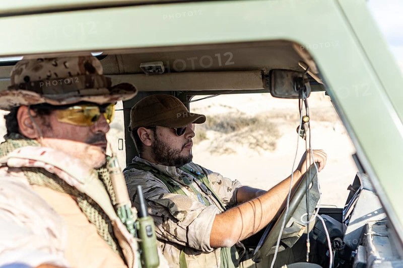 Two military soldiers in desert camouflage uniforms inside a vehicle. The driver is focused on the road while the passenger scans the surroundings, reflecting intense concentration
