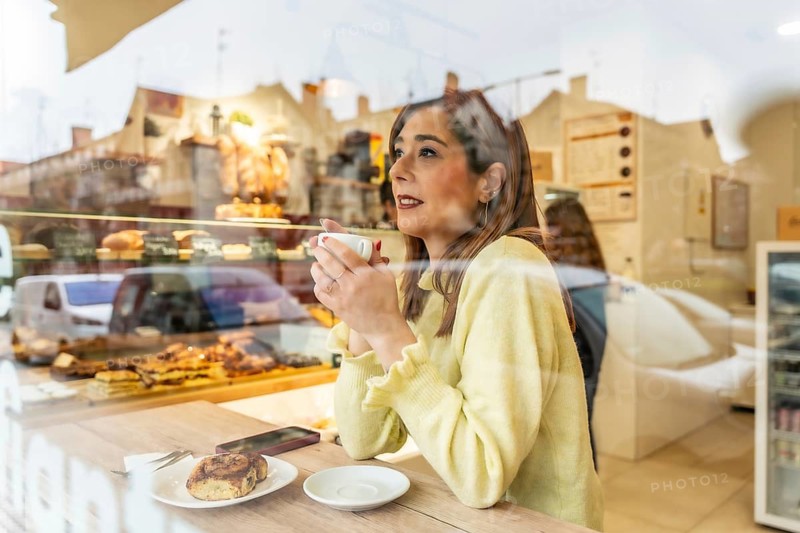 Young woman happily drinking coffee and looking away while sitting at a wooden table with a pastry, visible through the reflecting glass of a city bakery window