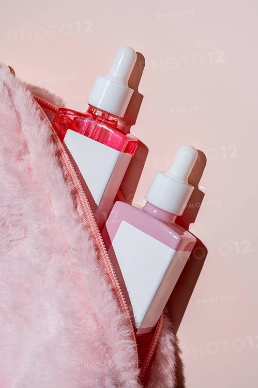 Two skincare serum bottles with dropper tops peek from a pink fur cosmetic bag. The pastel theme and soft lighting create a luxurious and feminine aesthetic