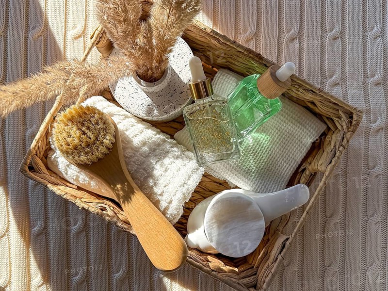 A top view of a woven basket containing spa essentials such as a natural brush, cosmetics, luxury oils, soft towels, and a marble dish. Set on a cozy, light textured knit fabric in the bathroom