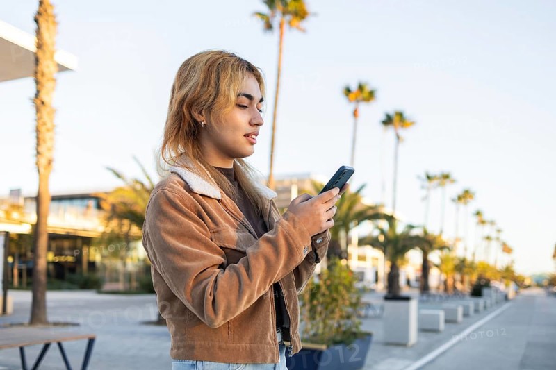 Mixed-race woman in casual jacket using a smartphone outdoors She appears to be busy with the device, with a background of palm trees in a beach setting