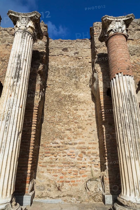Low angle view of old terracotta brick and stone wall remnants framed by marble columns with carved details at the ancient ruins of Pompeii, Campania region, Italy