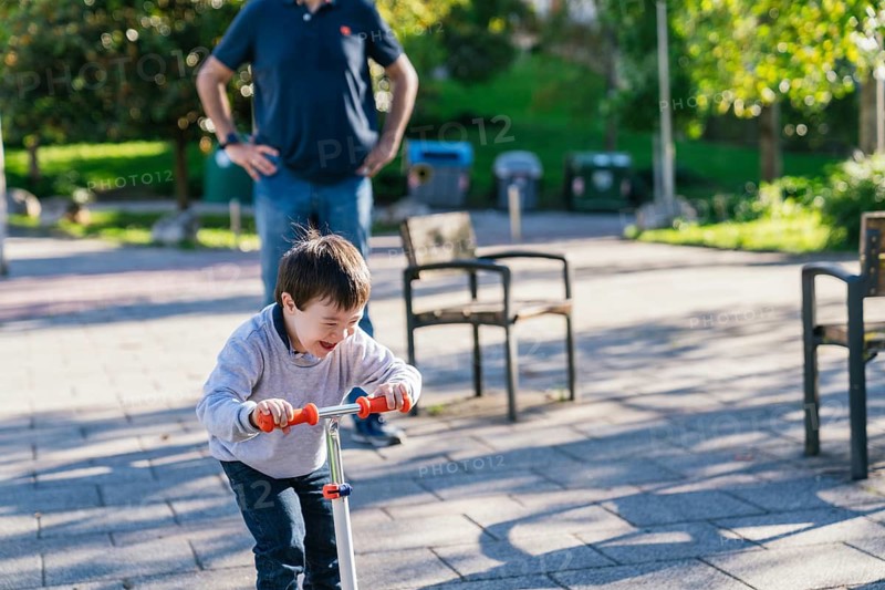 A cheerful child with Down syndrome rides a scooter through a sunlit park, laughing as his father watches nearby. The scene captures a moment of joy, freedom, and family bonding