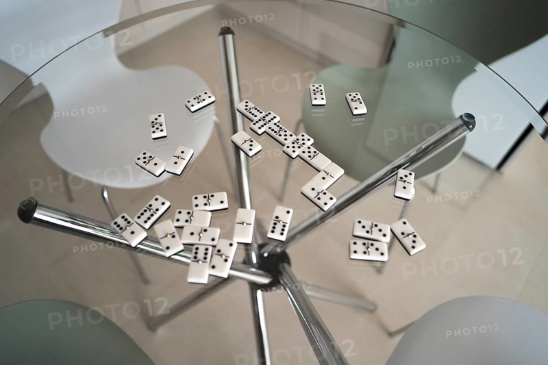 A top-down view of domino tiles scattered across a clear glass table. The modern setting features sleek, minimalist chairs, creating an inviting and elegant atmosphere