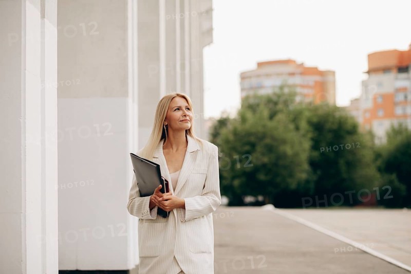 A businesswoman in a white suit holds a folder while walking on a city street. Urban buildings and lush trees create a balanced backdrop, reflecting professionalism