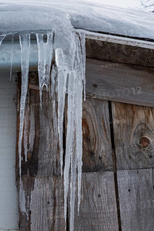 Long icicles hanging from snow and ice covered roof edge of old wood plank cladded building in winter, Quebec, Canada