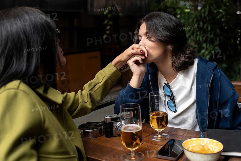 A lesbian couple enjoys a tender moment at a café, where one woman gently kisses the other's hand. Glasses of beer and snacks are on the table, creating a warm atmosphere