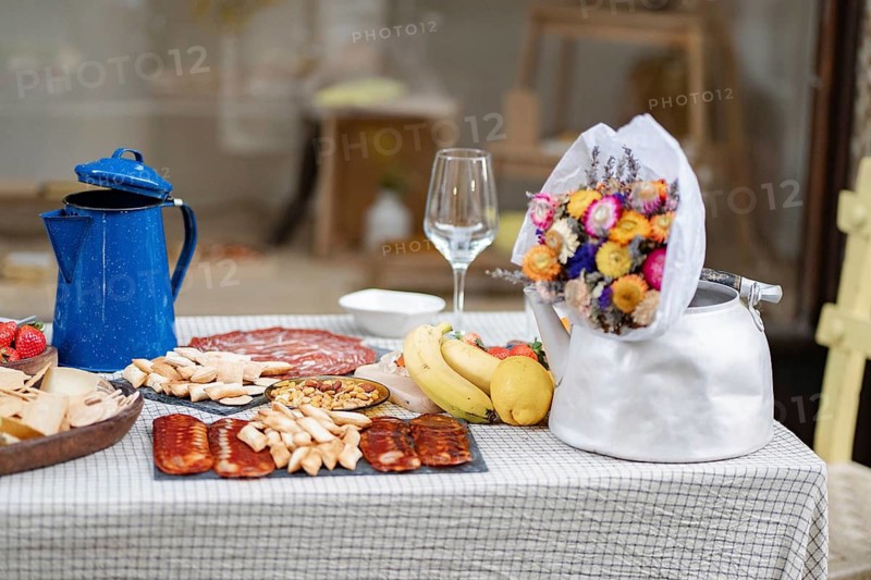 A rustic picnic setup on a checkered tablecloth, featuring dried flowers in a kettle, vibrant fruits, assorted cheeses, cured meats, and a blue coffee pot