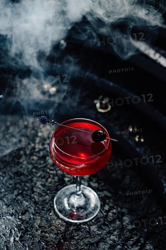A deep red berry infused gin cocktail with floral notes and sparkling wine splashes, elegantly garnished with a dark cherry. Set against a smoky backdrop, creating a dramatic and moody aesthetic