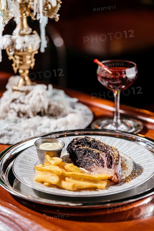 Elegant blue cabbage steak served with crispy fries and creamy sauce on a decorative plate. A sophisticated setting with a cocktail nearby enhances the gourmet experience