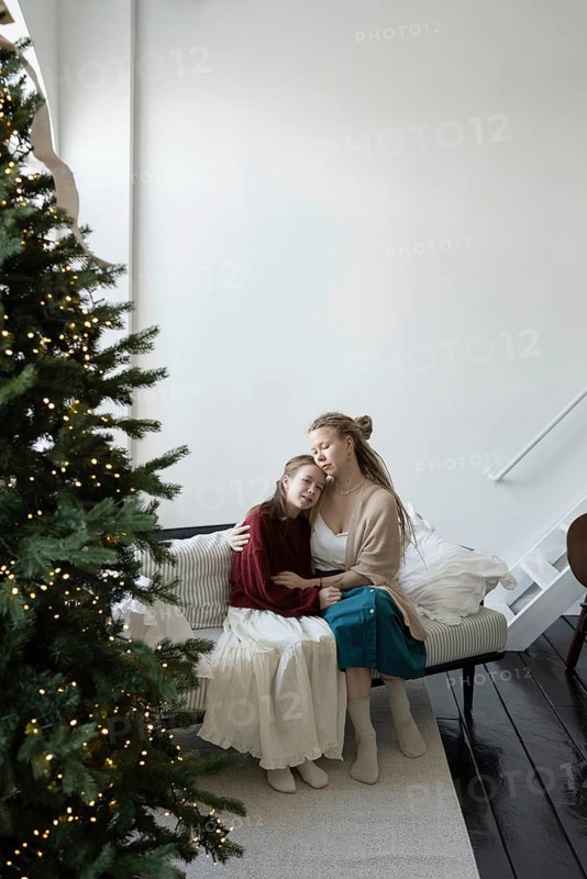 A mother and daughter share a tender moment, embracing on a cozy sofa next to a decorated Christmas tree The warm ambiance captures the essence of family love and holiday spirit