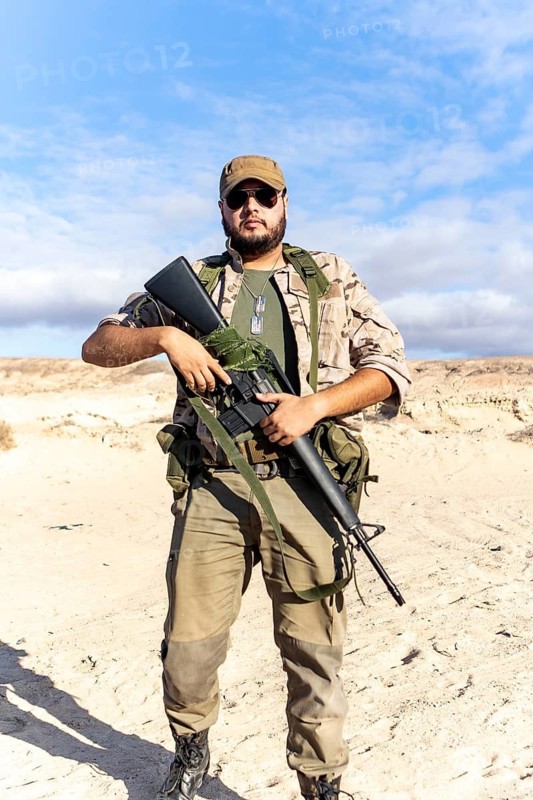 A soldier in camouflage holds a rifle in a desert setting. The clear sky adds contrast. The focus is on military readiness, tactical gear, and desert operations