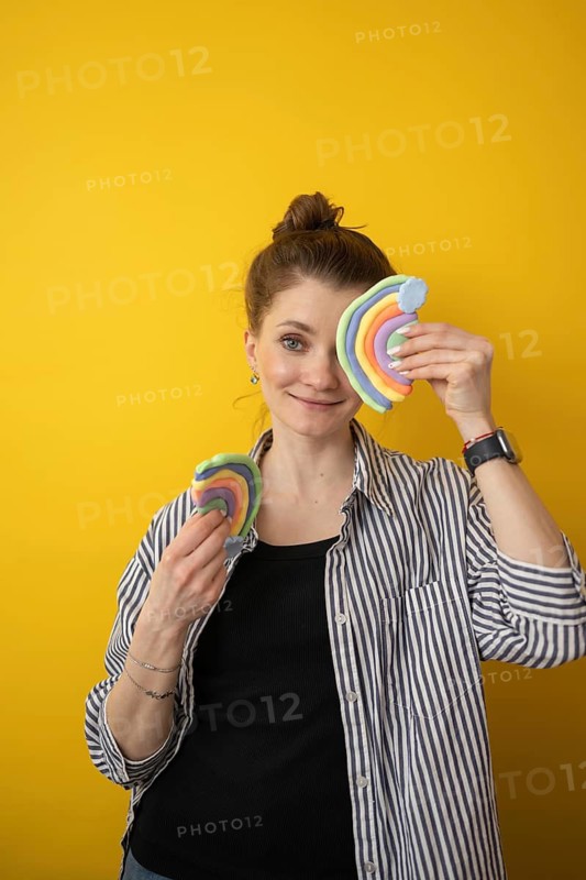 A woman joyfully holds two colorful clay rainbows against a vibrant yellow background. This cheerful scene captures the essence of creative expression and playful learning