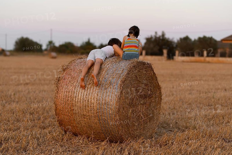 Back view of unrecognizable young boy in shorts lies atop a large hay bale while the girl, dressed in a striped shirt, sits observing the horizon. They are surrounded by a golden field under a dusky sky