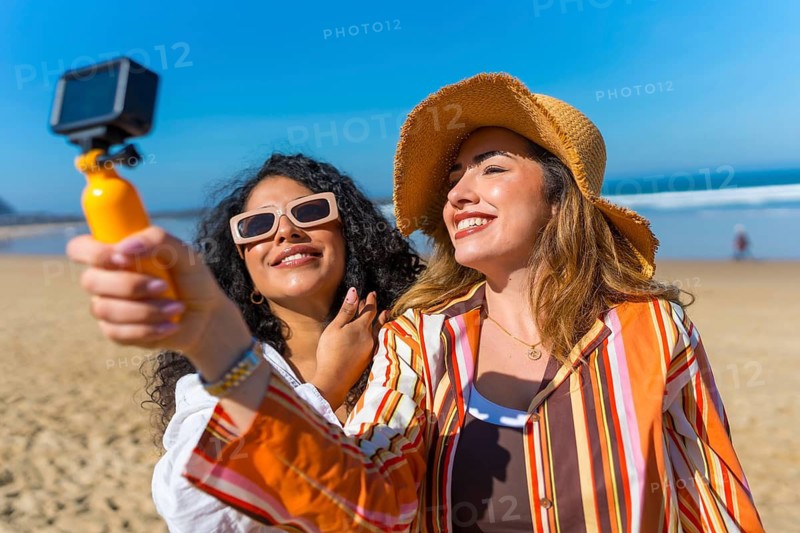 Two smiling women friends enjoying a sunny beach day, holding an action camera on a selfie stick and recording moments of their summer vacation together