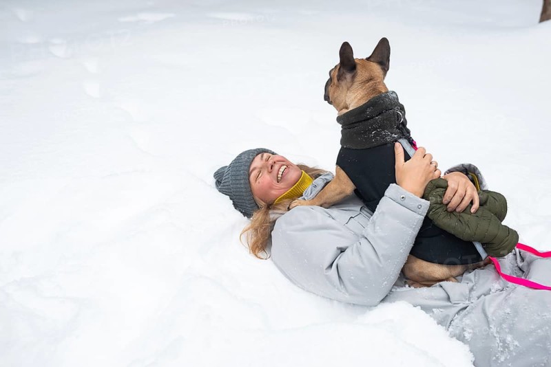 A person in a gray winter coat and beanie joyfully playing in the snow with a dressed-up dog. The snowy setting captures the essence of winter fun and companionship