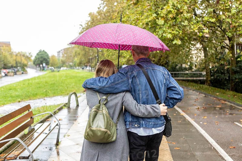 A couple with intellectual disabilities walks arm in arm under a pink polka dot umbrella on a rainy day. They stroll along a wet path, surrounded by lush green trees and a peaceful urban setting