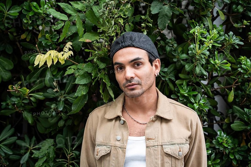 A gay man in casual clothing stands against a lush, leafy background. He sports a chain necklace and earring, conveying a relaxed and stylish vibe in natural surroundings