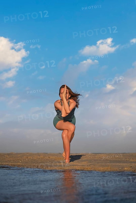 Woman stands on one leg with the other wrapped, arms crossed, practicing yoga on the beach at sunset