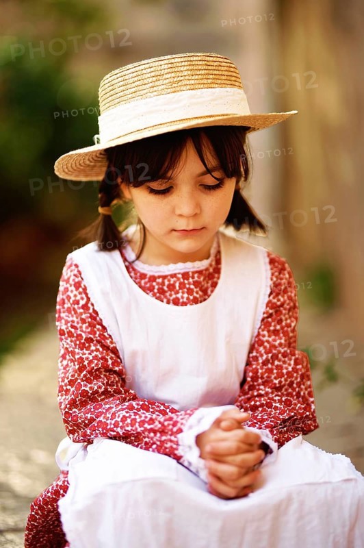 A young girl with brown hair wearing a straw hat and a red vintage dress with a white apron. She is seated with her hands folded, conveying a contemplative mood