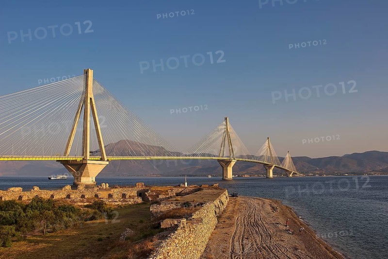 A vast bridge across the sea with beach facilities and ruins in the foreground against a mountain backdrop, Rio-Antirrio cable bridge, Rio fortress, Patras, Peloponnese, Greece