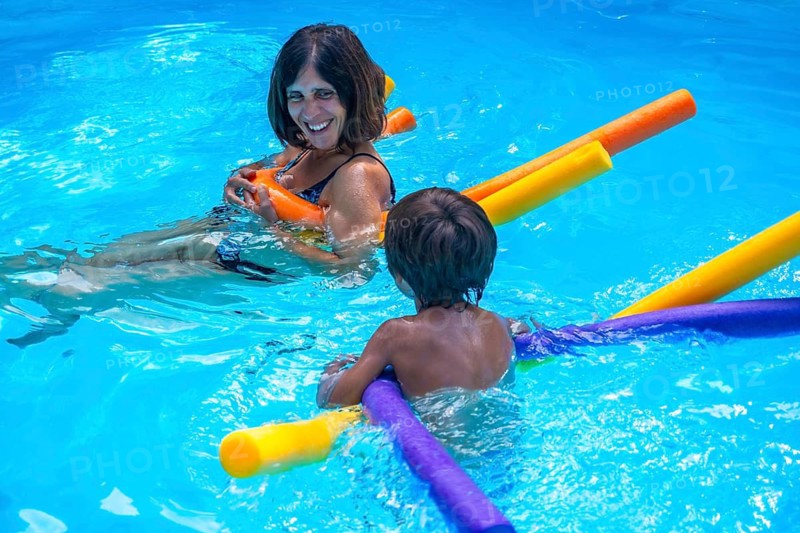 Happy mother smiling, holding a pool noodle and looking at her young son, who is also playing with colorful pool noodles in bright blue swimming pool water during summer