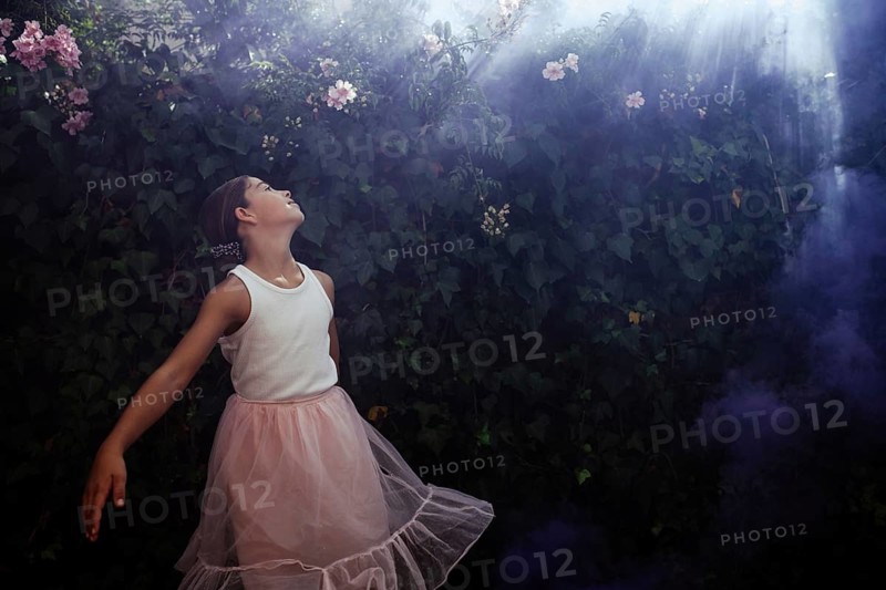 A teenage girl in a white blouse and pink skirt gazes enchanted at a lush garden, with sunlight filtering through the leafy background and a light smoke drifting in. The atmosphere is magical