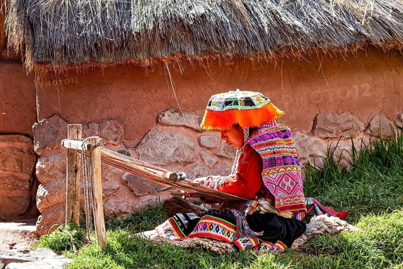 Person in traditional, colorful clothing weaving in front of a thatched hut in a meadow, elderly traditionally dressed lady from Peru weaving a cloth