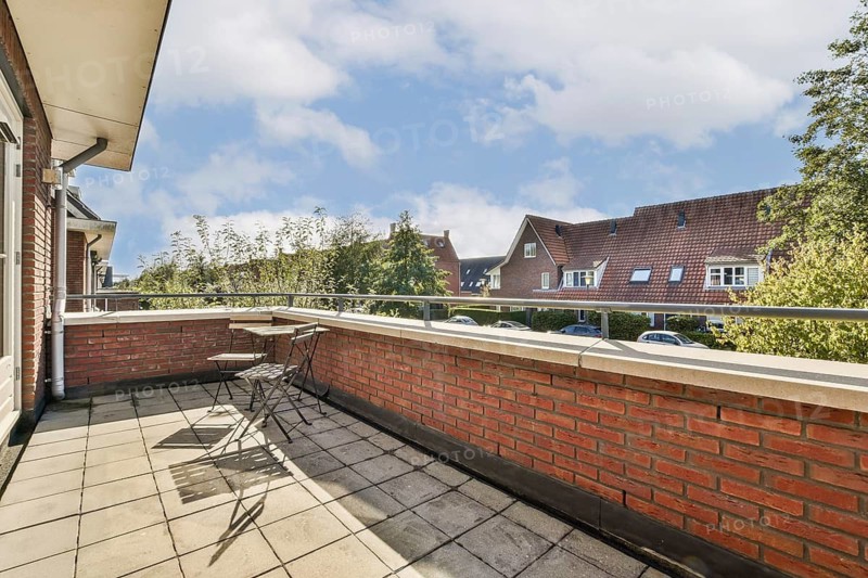A sunny balcony with two chairs and a table overlooks a quaint neighborhood with brick houses. The clear blue sky enhances the peaceful, inviting atmosphere of the scene