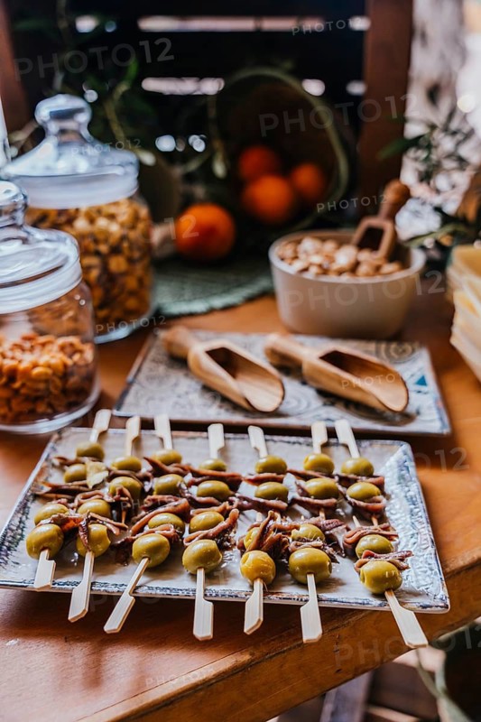 A tray of appetizer skewers with green olives and anchovies is beautifully arranged on a rustic wooden table, surrounded by snacks, fruits, and natural textures in a cozy setting