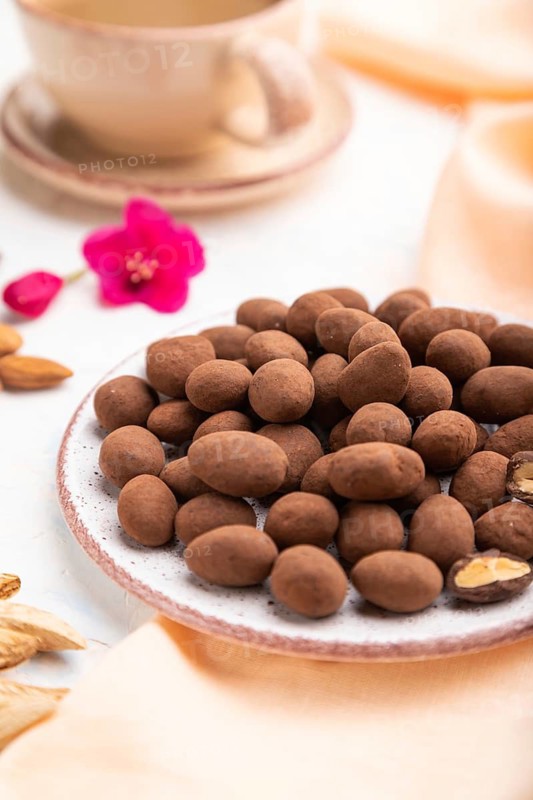Almond in chocolate dragees on ceramic plate and a cup of coffee on white concrete background and orange linen textile. Side view, close up, selective focus