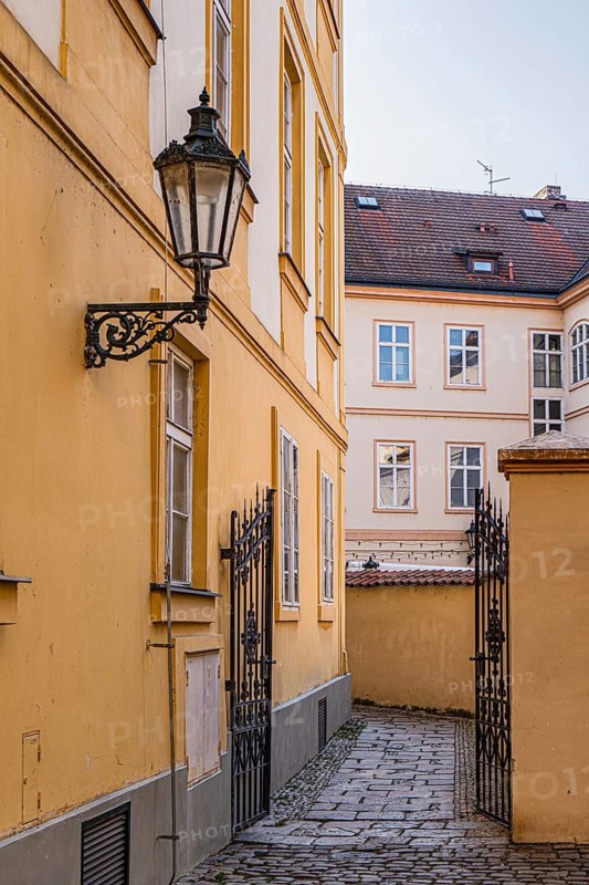 Quaint alleyway flanked by yellow buildings in Prague, featuring an ornate lantern and wrought iron gate. Cobblestone path adds vintage charm to the picturesque European scene