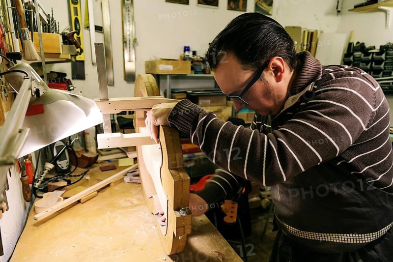 A luthier carefully crafts a bass guitar in a workshop filled with specialized tools, focusing intently on shaping the guitar body with precision
