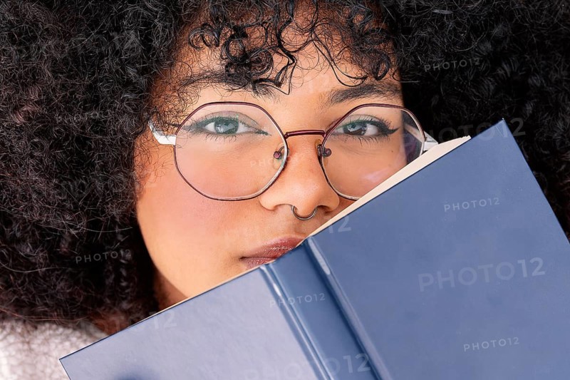 A latin woman with curly hair peers over a blue book, wearing large round glasses. Her expression is focused, highlighting a sense of curiosity and contemplation