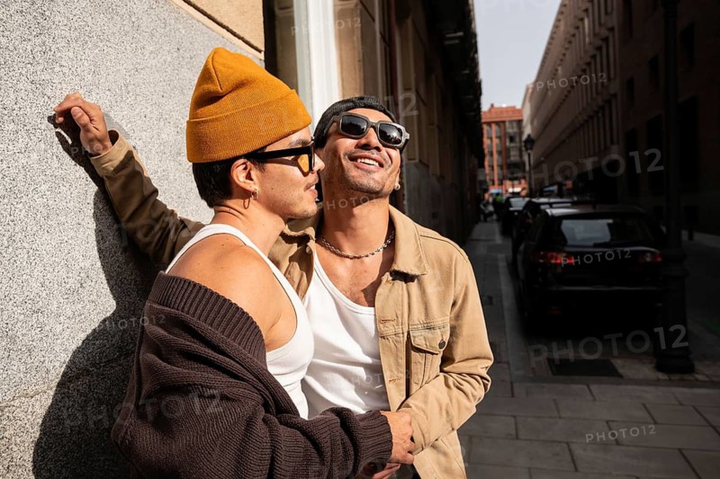 A gay couple in fashionable attire enjoys a sunny day in an urban setting. They smile warmly, exuding happiness and connection, capturing the essence of modern love