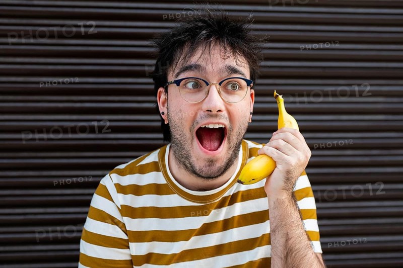 A man with glasses and a striped shirt humorously uses a banana as a phone. He displays a surprised expression, standing against a dark, striped background