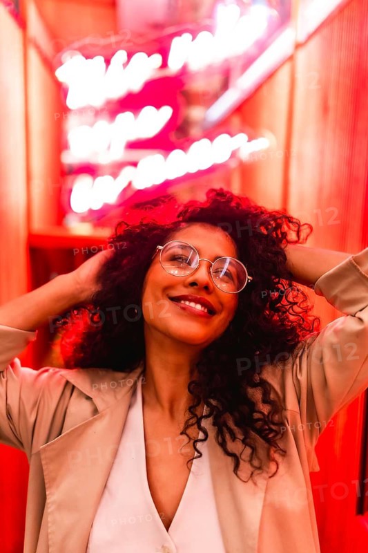 Vertical portrait of a sensual latin woman with hands on hair smiling indoors illuminated with red neon lights