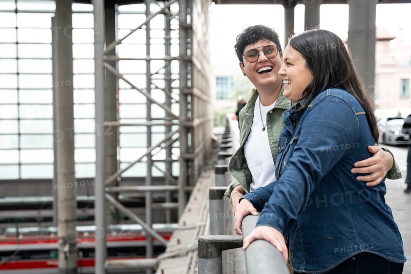 A happy lesbian couple shares a joyful moment, leaning on a railing in a train station. Both women are smiling and embracing, exuding warmth and affection